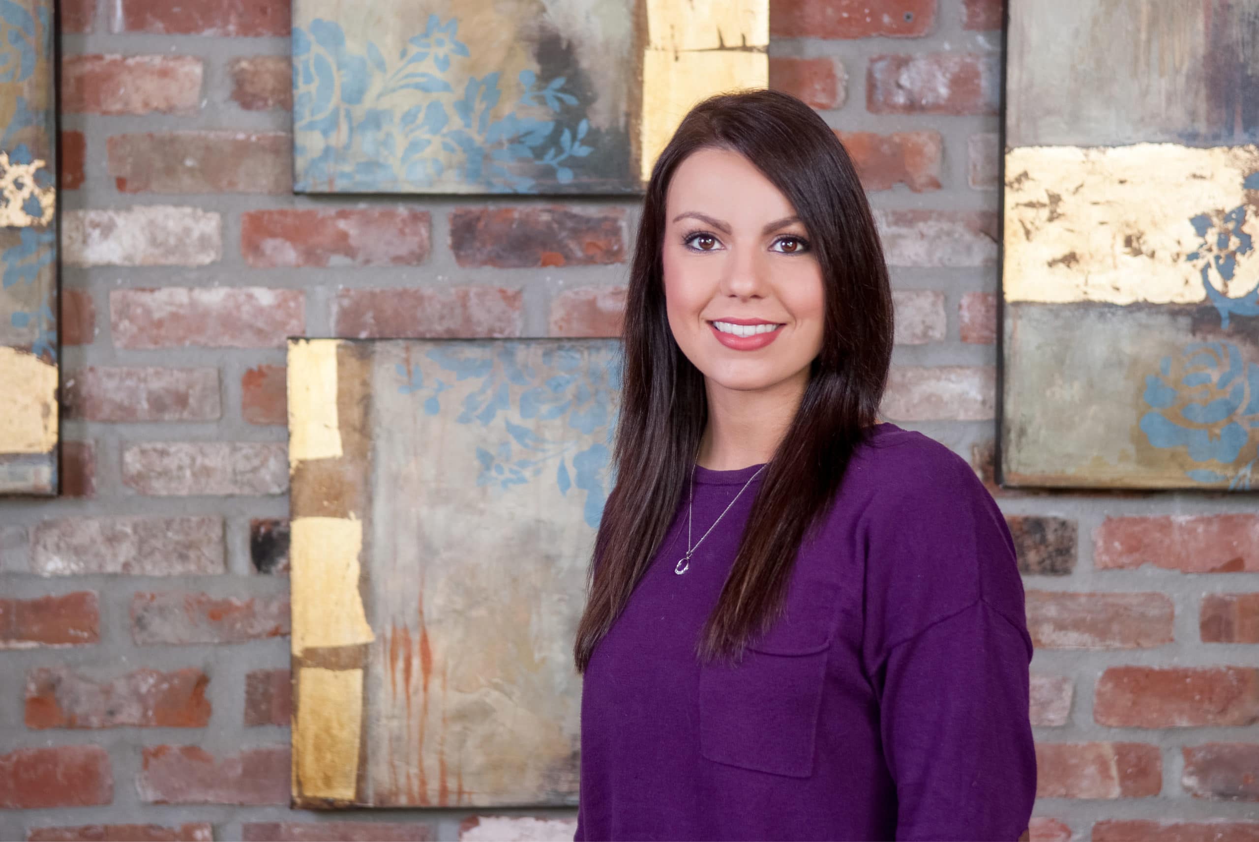 a woman in a purple shirt stands in front of a brick wall