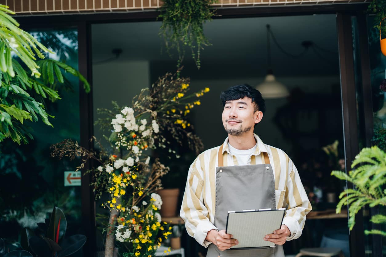 Portrait of confident young Asian male florist, owner of small business flower shop, standing in front of flower shop with clipboard on hand. He is looking away with smile. Small business concept