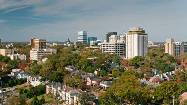 Aerial shot of Columbia, South Carolina from over the Five Points neighborhood, looking across the rooftops and the University of South Carolina campus towards downtown office towers and the state house.